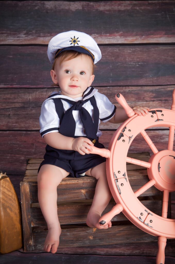 A two-year old boy in a cute sailor suit sitting next to a ship's steering wheel