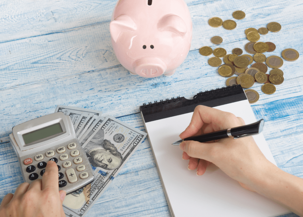 Woman's right hand is writing a budget on a pad of paper while her left hand is using a calculator. Coins are spread on the table next to a piggy bank.