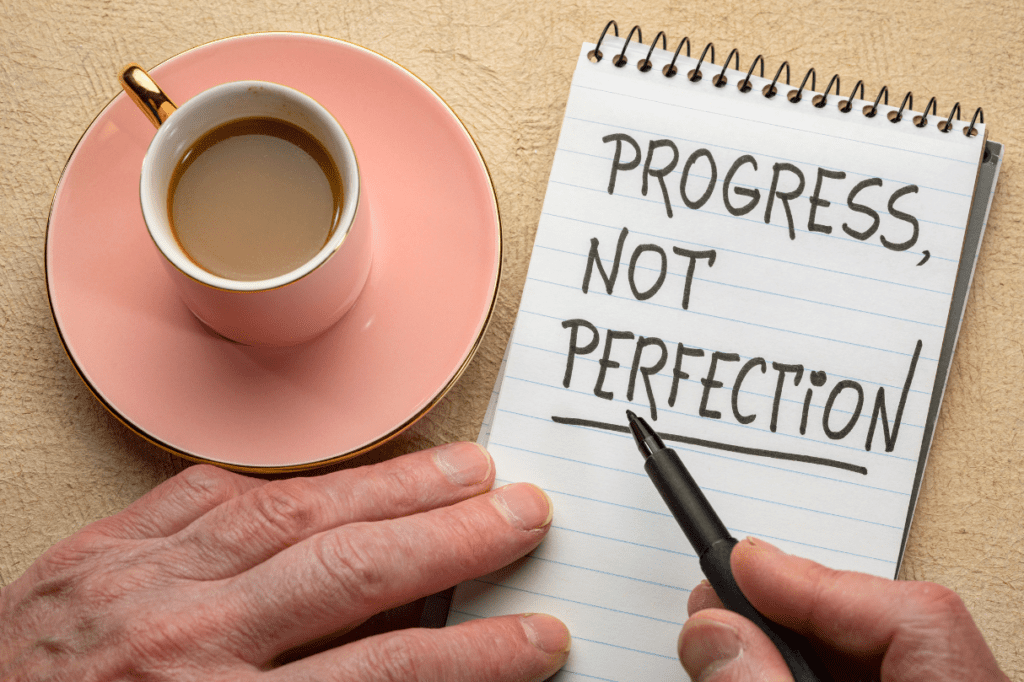 Pair of hands writing 'progress not perfection' on a note pad with a cup of coffee in a cup and saucer nearby