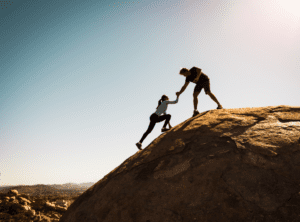 Silhouettes of 2 people climbing a mountain. 1 person has an outstretched hand to help the other person up.