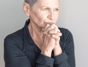 Older woman is sitting against a gray background with her hands clasped. She looks worried.